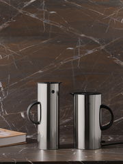 Zwei EM77 Wasserkocher aus Edelstahl mit schwarzem Griff, frontal auf einer Marmoroberfläche vor einer marmorierten Wand platziert.