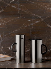 Zwei EM77 Wasserkocher aus Edelstahl mit schwarzem Griff, frontal auf einer Marmoroberfläche vor einer marmorierten Wand platziert.