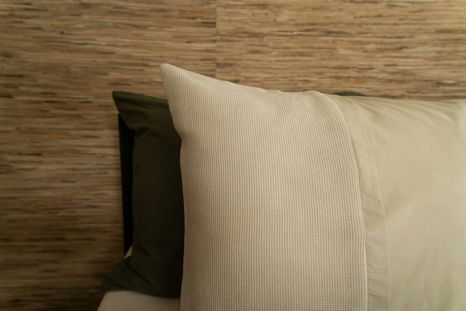 Nahaufnahme von Bettwäsche in Beige und Olivgrün vor einer Holzwand.