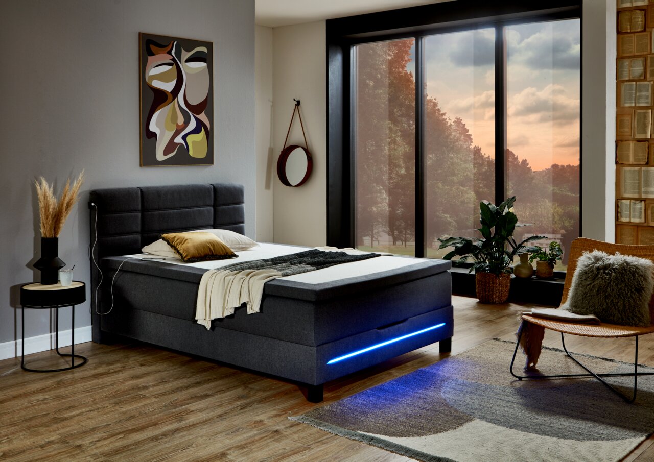 Modernes Boxspringbett in einem stilvoll eingerichteten Schlafzimmer mit Blick auf große Fenster und eine gemütliche Atmosphäre, seitliche Perspektive.