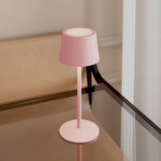 Rosa, minimalistische Tischleuchte mit zylindrischem Schirm und rundem Fuß auf einem Glastisch neben einer beigefarbenen Sitzbank, fotografiert aus leicht erhöhter schräger Seitenperspektive.