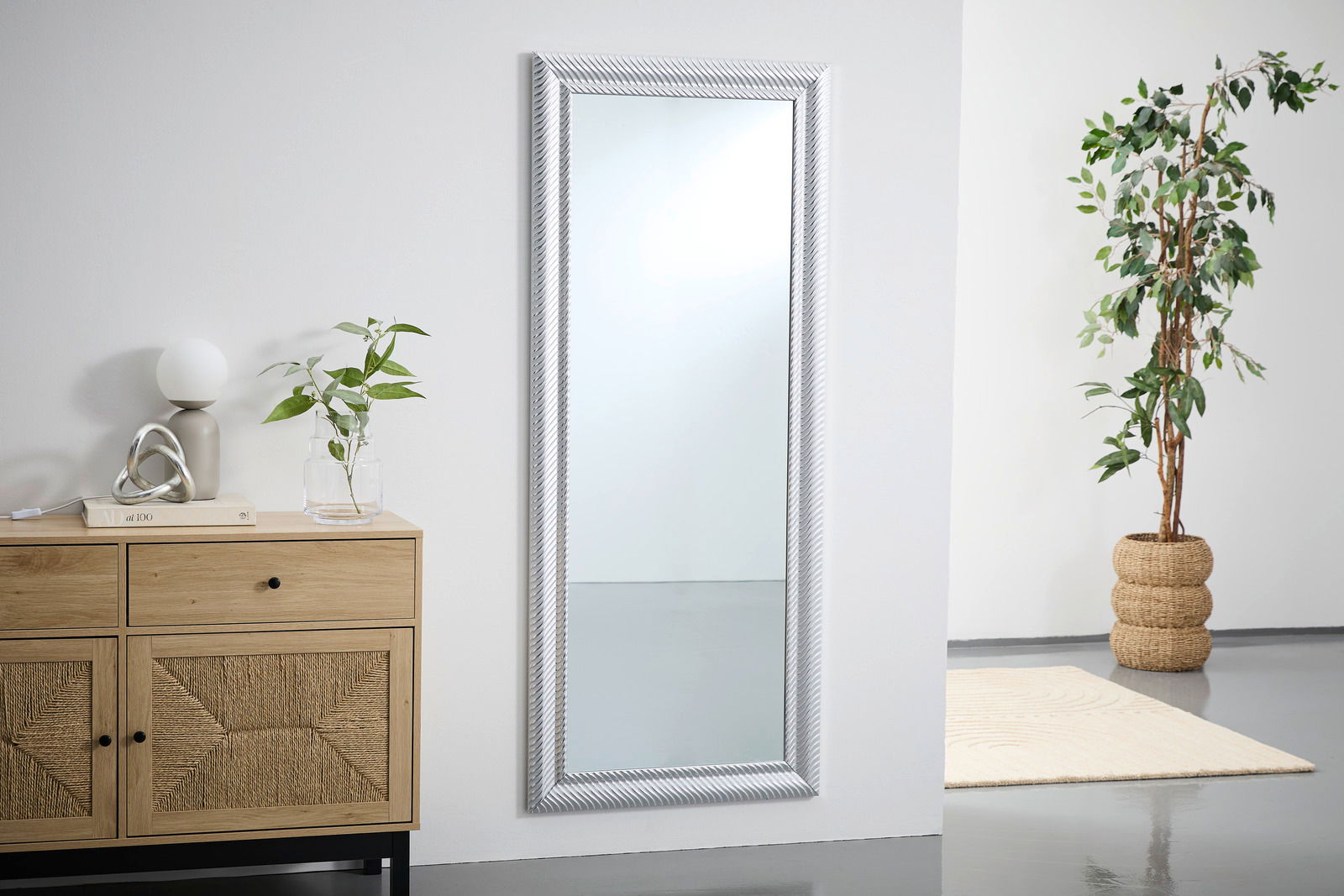 Großer rechteckiger Spiegel mit silbernem Rahmen, an einer weißen Wand lehnend, neben einer Kommode und einer Pflanze, aus seitlicher Perspektive.
