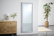Großer rechteckiger Spiegel mit silbernem Rahmen, an einer weißen Wand lehnend, neben einer Kommode und einer Pflanze, aus seitlicher Perspektive.