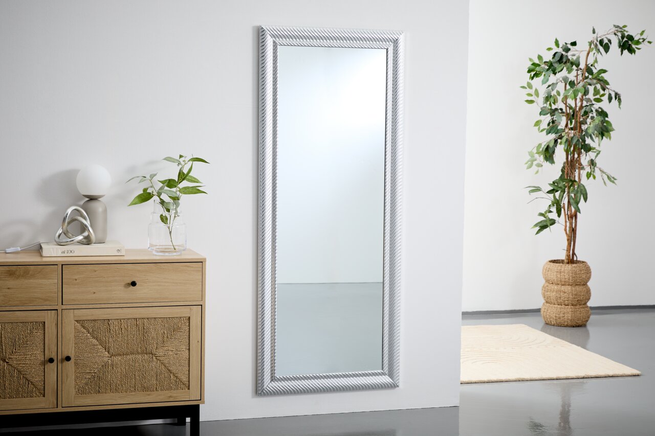 Großer rechteckiger Spiegel mit silbernem Rahmen, an einer weißen Wand lehnend, neben einer Kommode und einer Pflanze, aus seitlicher Perspektive.