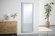 Großer rechteckiger Spiegel mit silbernem Rahmen, an einer weißen Wand lehnend, neben einer Kommode und einer Pflanze, aus seitlicher Perspektive.