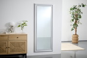 Großer rechteckiger Spiegel mit silbernem Rahmen, an einer weißen Wand lehnend, neben einer Kommode und einer Pflanze, aus seitlicher Perspektive.