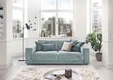 Zweiersofa in einem hellen Wohnzimmer mit großen Fenstern, dekoriert mit Kissen in verschiedenen Blautönen, aus der Frontalperspektive.