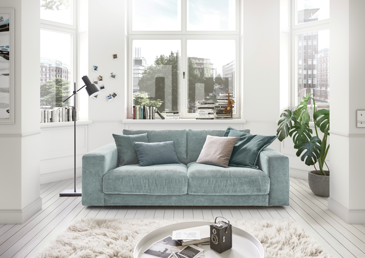 Lounge Handmade Sofa 2-Sitzer LH-SAMBIA Zweiersofa in einem hellen Wohnzimmer mit großen Fenstern, dekoriert mit Kissen in verschiedenen Blautönen, aus der Frontalperspektive.