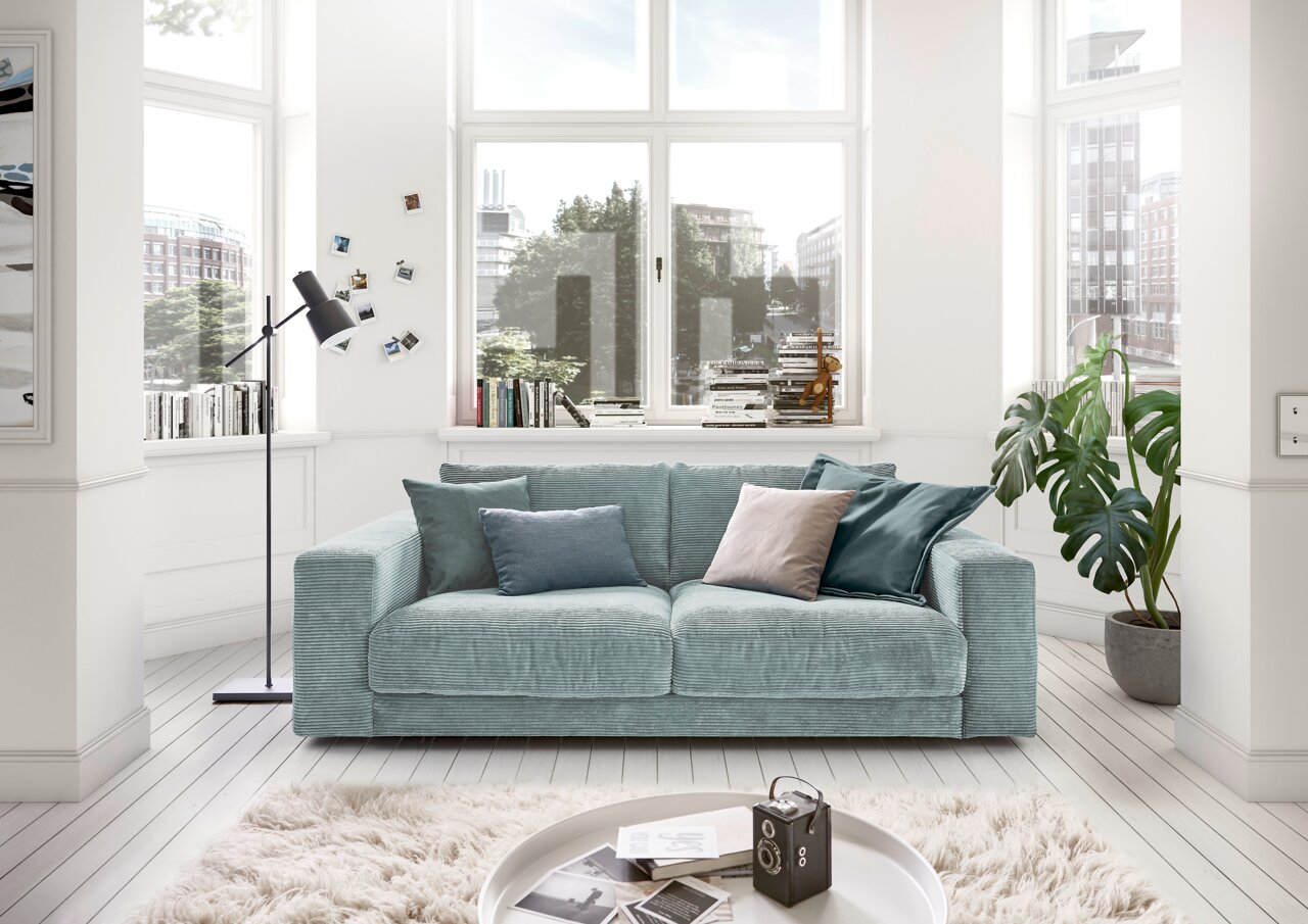 Zweiersofa in einem hellen Wohnzimmer mit großen Fenstern, dekoriert mit Kissen in verschiedenen Blautönen, aus der Frontalperspektive.