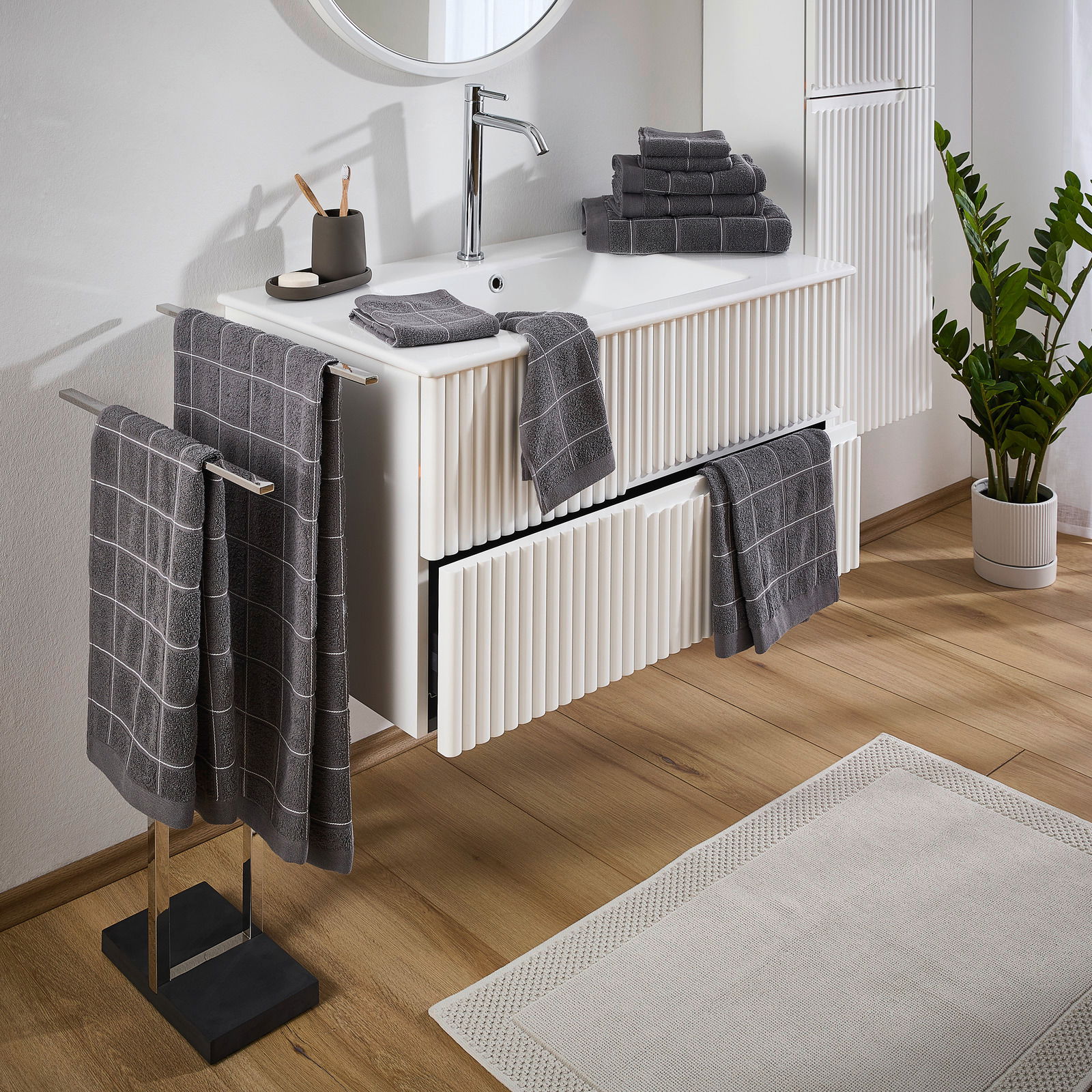 Badezimmer mit grauem Frottierset, bestehend aus Handtüchern mit Karomuster, auf einem weißen Waschtisch und Handtuchhalter. Perspektive von schräg oben.