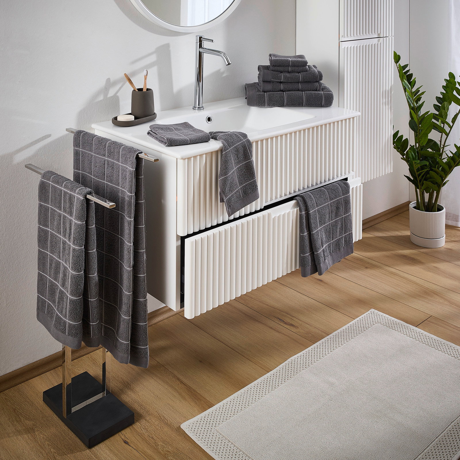 Badezimmer mit grauem Frottierset, bestehend aus Handtüchern mit Karomuster, auf einem weißen Waschtisch und Handtuchhalter. Perspektive von schräg oben.