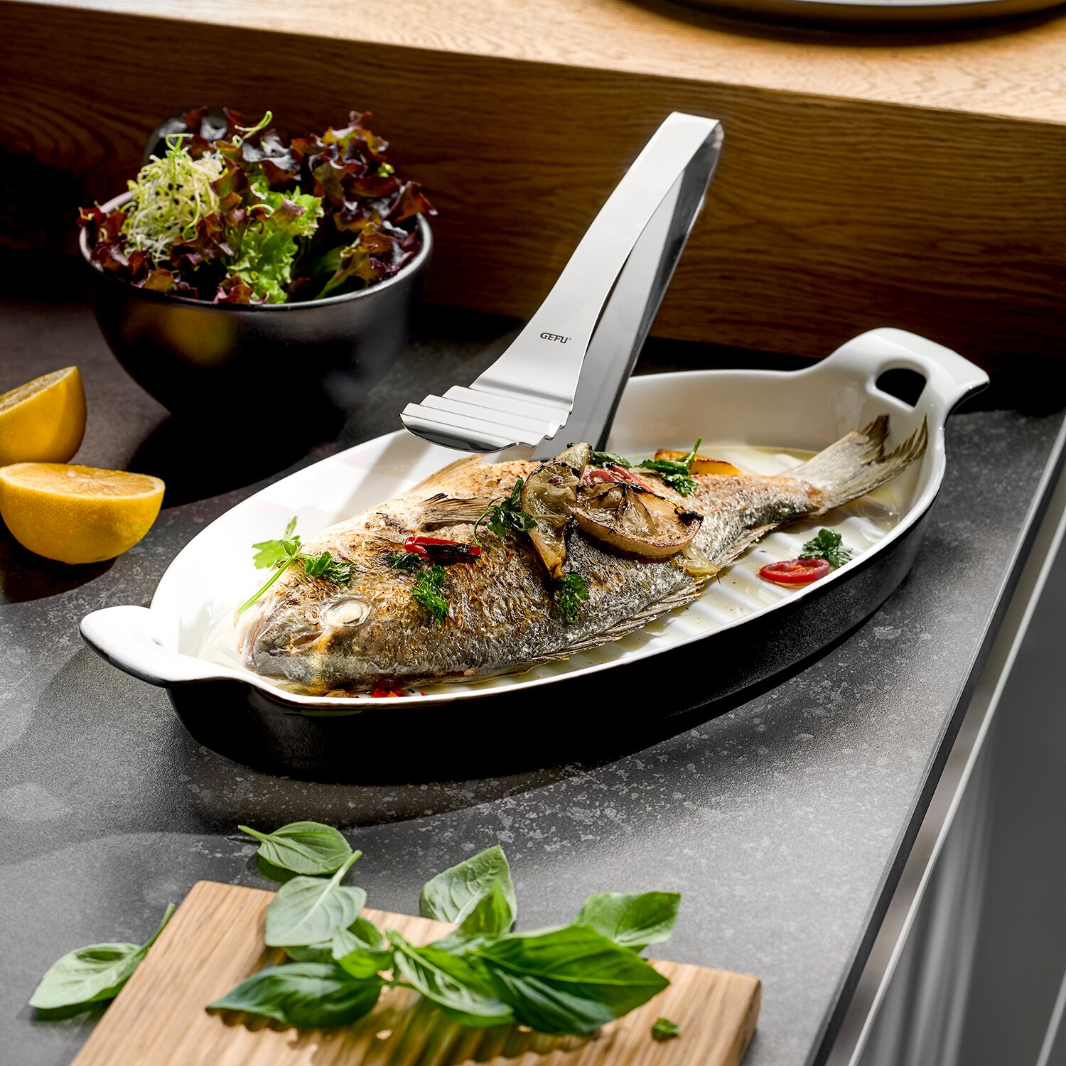GEFU Servierplatte 40cm mit Zange PRESENTO Edelstahl-Fischgrätenpinzette greift eine gebratene ganze Dorade in einer weißen Ofenform, Küchen-Setting mit Zitrone und Salat, perspektivisch in leichter Draufsicht (Schrägansicht von vorn)