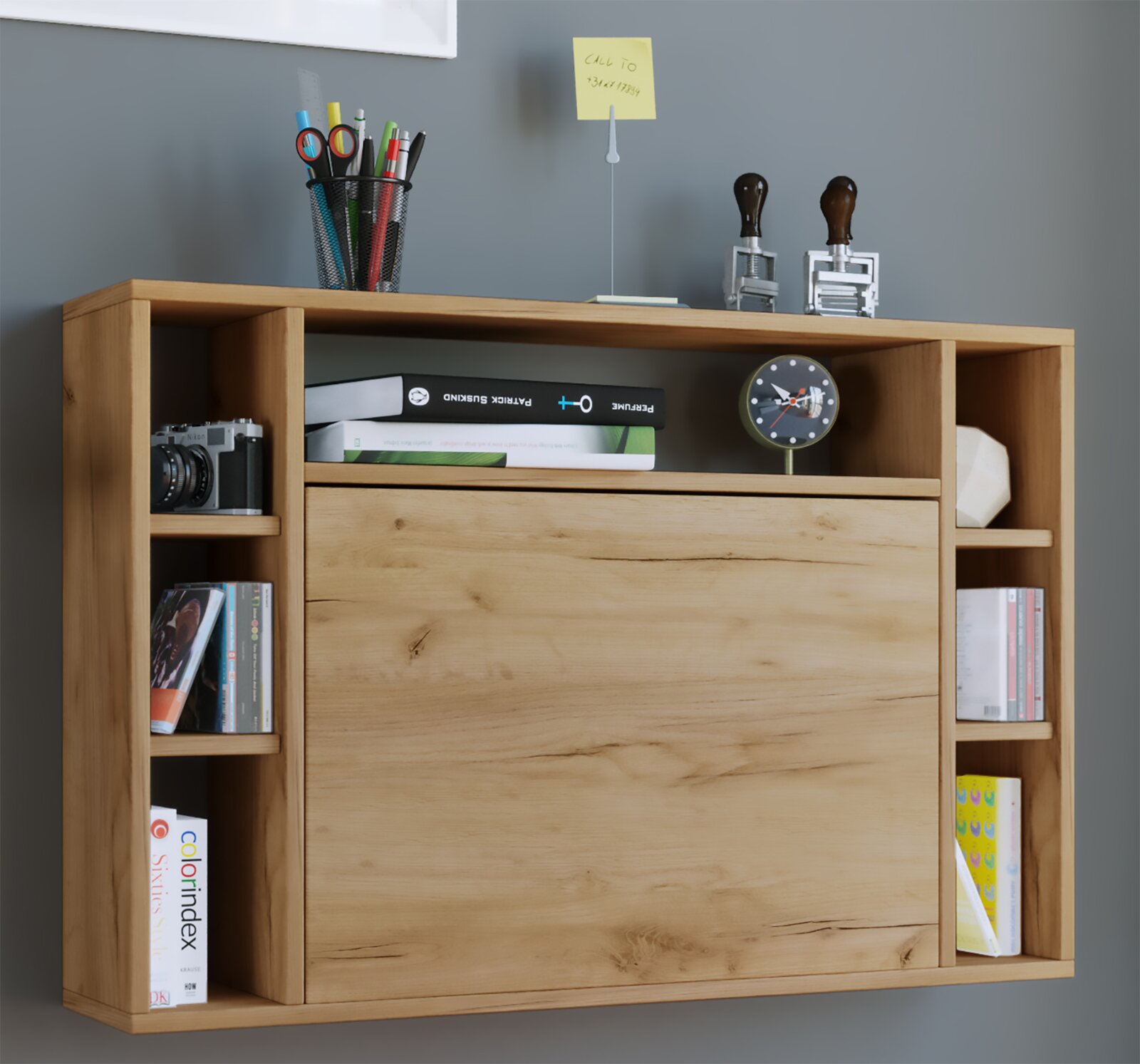 Wandhängendes Holzregal in Eiche-Optik mit großer Klapptür und seitlichen Fächern, dekoriert mit Büchern, Kamera und Stiftebecher, Frontansicht leicht von links.