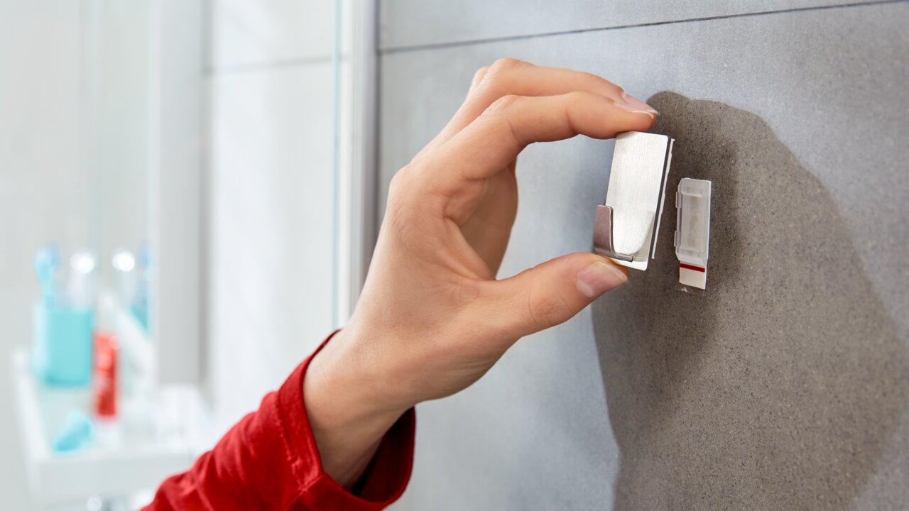 Nahaufnahme einer Hand, die einen tesa Powerstrip Waterproof Haken an einer Badezimmerwand anbringt.