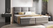 Modernes Boxspringbett in grauem Stoff mit hohem Kopfteil, dekoriert mit Kissen und Decke, seitliche Perspektive.