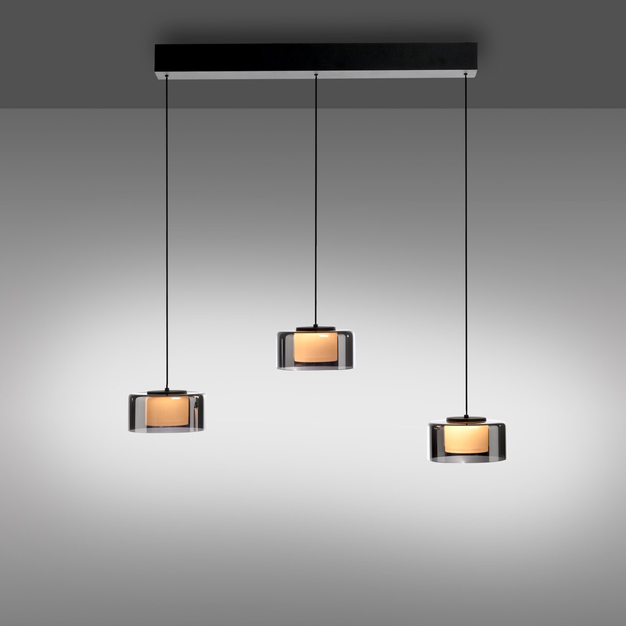Drei moderne Pendelleuchten RIKA mit zylindrischen, transparenten Schirmen und warmem Licht, von vorne betrachtet.