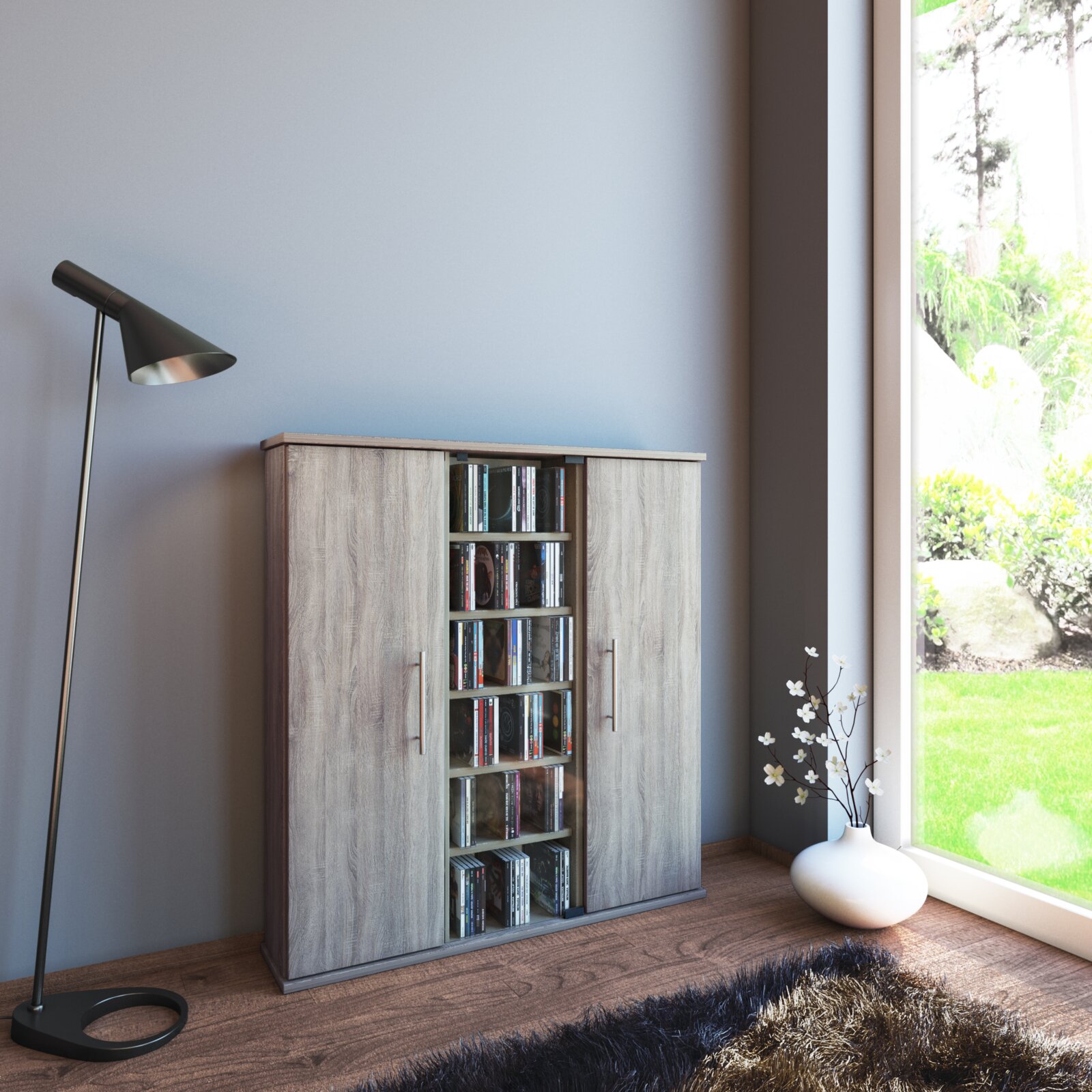 Schmaler Schrank in grauer Holzoptik mit zwei seitlichen Türen und mittlerem Regal mit CDs, in einem modernen Wohnraum neben Fenster und Stehlampe, perspektivisch frontal aus leicht linker Seitenansicht