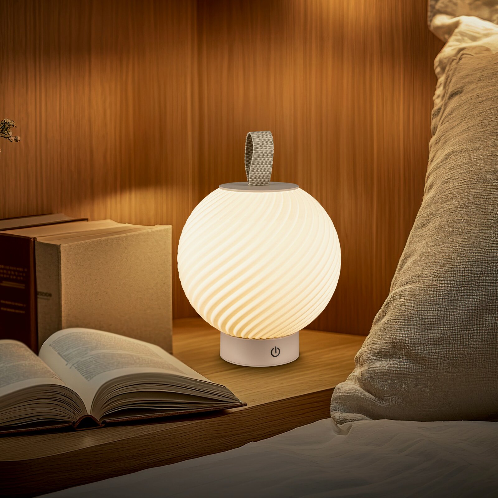 Leuchtende, kugelförmige LED-Nachttischlampe mit geriffelter Oberfläche und Trageschlaufe auf Holz-Nachttisch neben Bett und Buch, frontale Perspektive auf Augenhöhe