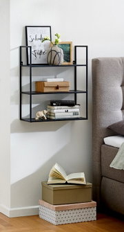 Schwarzes, wandmontiertes Metallregal mit drei Ablagen neben einem grauen Sofa, dekoriert mit Bilderrahmen, Vase, Büchern und Boxen, seitliche Perspektive im Wohnraum.