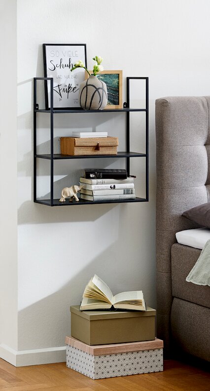 Zurbrüggen Wandregal Schwarzes, wandmontiertes Metallregal mit drei Ablagen neben einem grauen Sofa, dekoriert mit Bilderrahmen, Vase, Büchern und Boxen, seitliche Perspektive im Wohnraum.