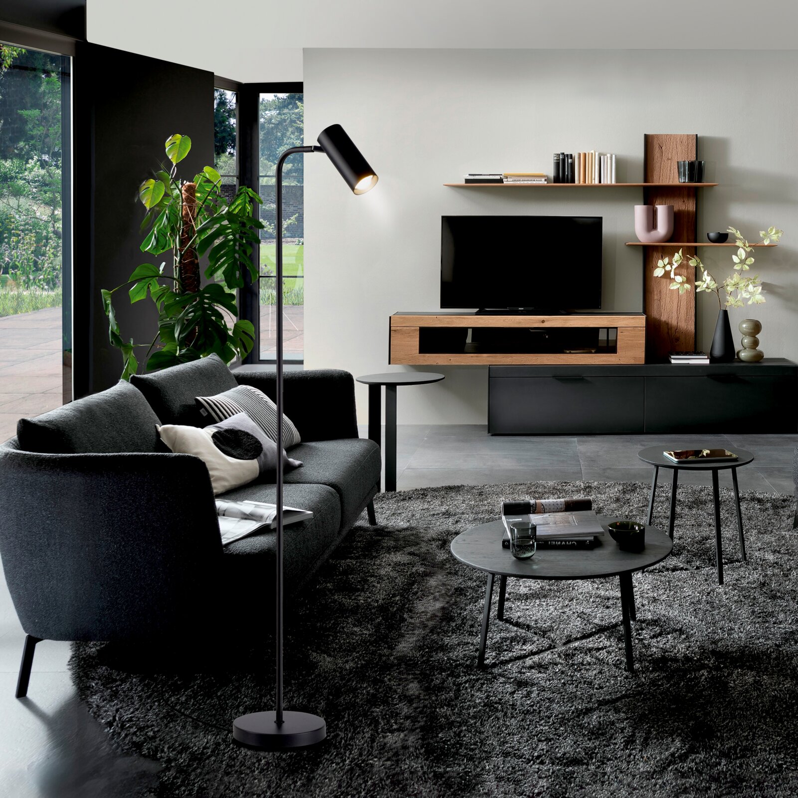 Modernes Wohnzimmer mit schwarzem Sofa, Stehlampe und TV-Wandmöbel aus Holz/Schwarz mit Regalen; Perspektive: frontale Raumansicht leicht von links.