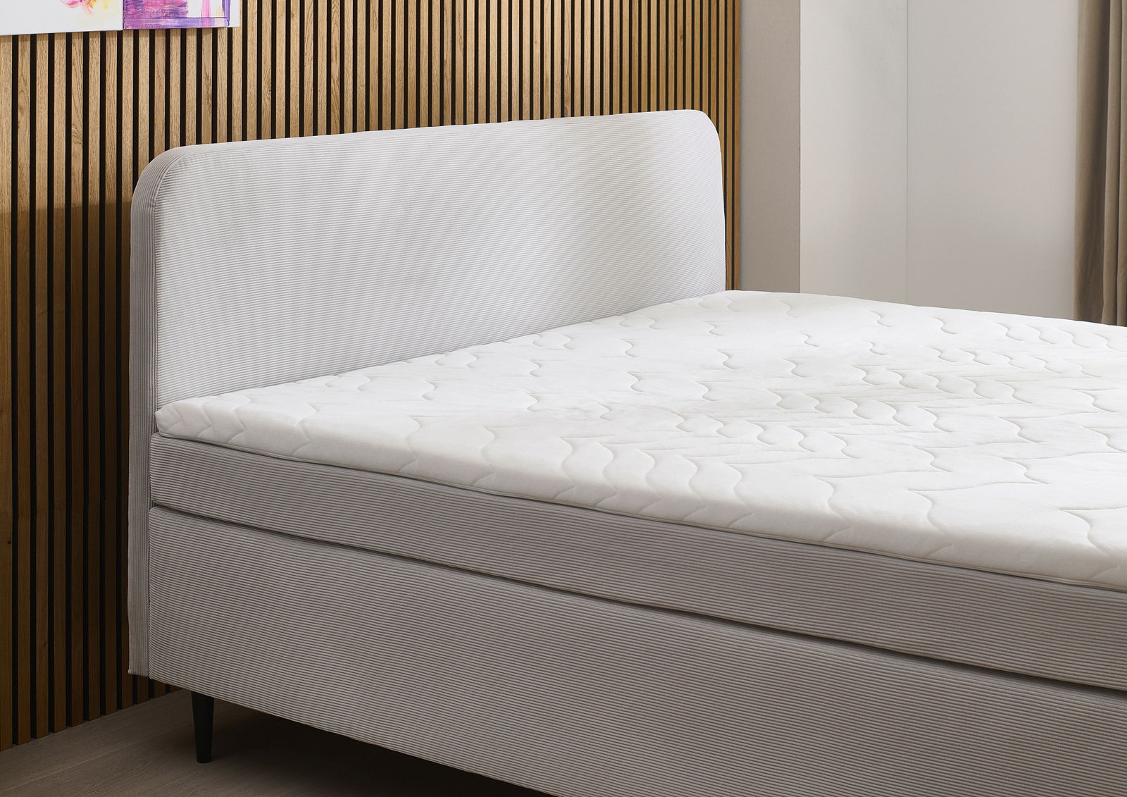 Seitliche Ansicht eines modernen Boxspringbetts mit gepolstertem Kopfteil und weißer Matratze vor einer Holzwand.