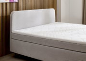 Seitliche Ansicht eines modernen Boxspringbetts mit gepolstertem Kopfteil und weißer Matratze vor einer Holzwand.