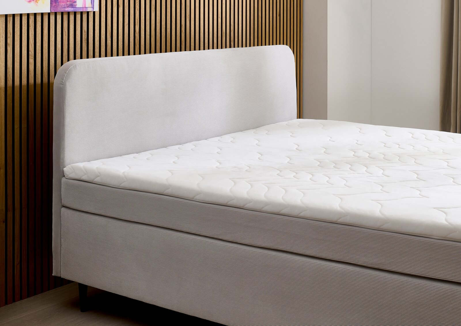 Zijaanzicht van een modern boxspringbed met een gestoffeerd hoofdeinde en een wit matras tegen een houten wand.