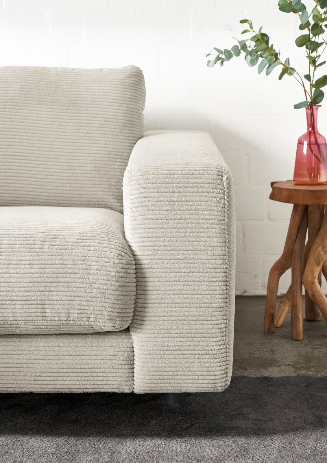 Nahaufnahme eines beigen Longchairs mit geripptem Stoffbezug, von der Seite betrachtet. Daneben steht ein kleiner Holztisch mit einer roten Vase und grünen Zweigen.