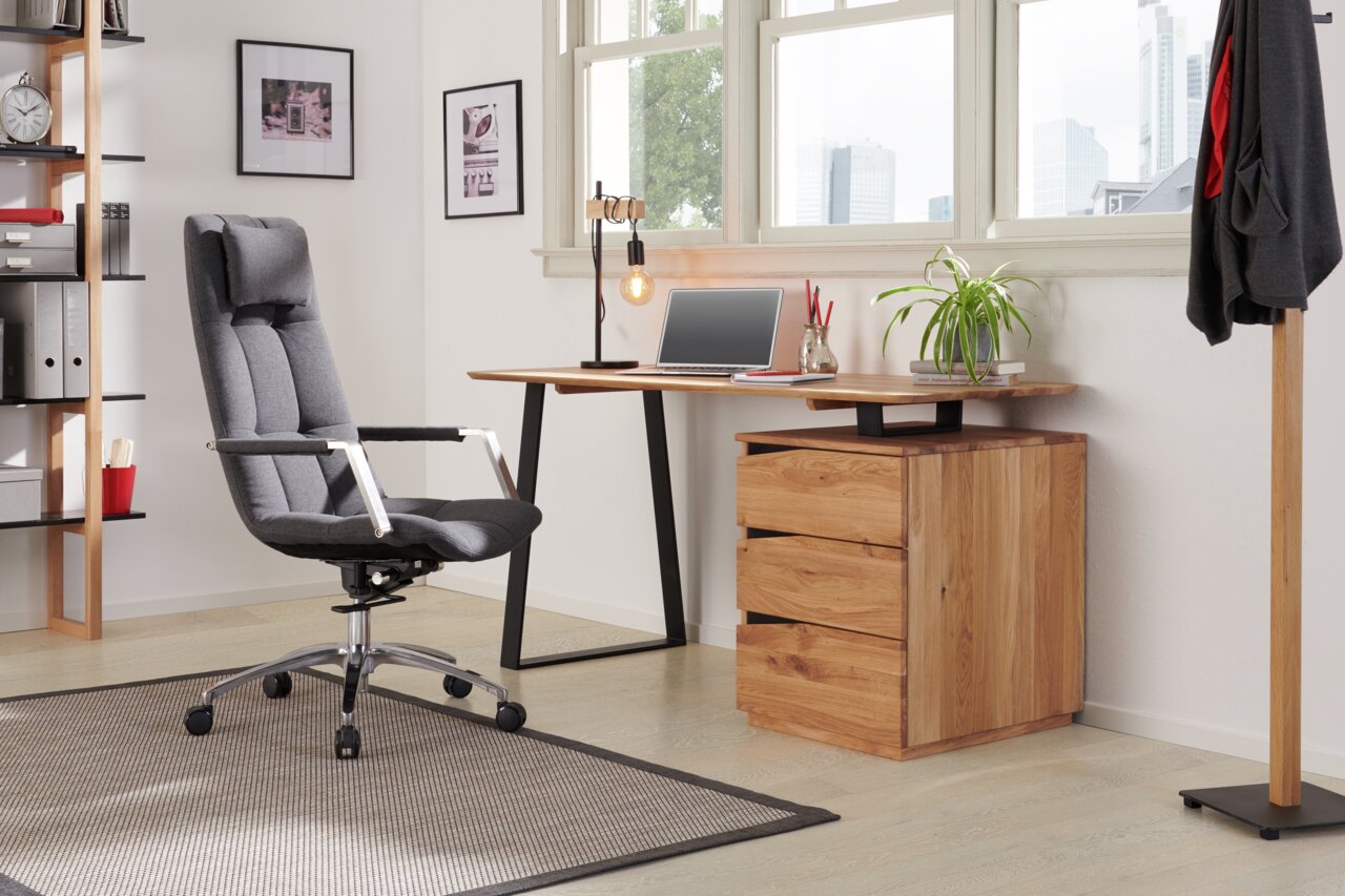 Moderner Schreibtisch mit Holzoberfläche und schwarzen Metallbeinen, seitlich von einem ergonomischen Bürostuhl und einem Holzschrank mit Schubladen flankiert, in einem hellen Raum mit großen Fenstern.