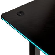 Detailansicht eines schwarzen Gaming-Schreibtischs mit Carbon-Optik, integrierter blauer LED-Kantenbeleuchtung und Kabeldurchlass in der Tischplatte, fotografiert aus schräger Draufsicht.