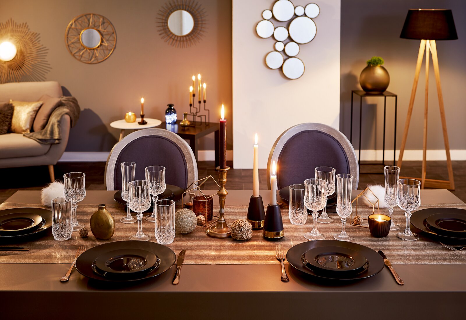 Festlich gedeckter Esstisch mit schwarzen Tellern, Kristallgläsern und Kerzen auf goldenem Tischläufer in modernem Wohnzimmer, frontal aus Augenhöhe aufgenommen