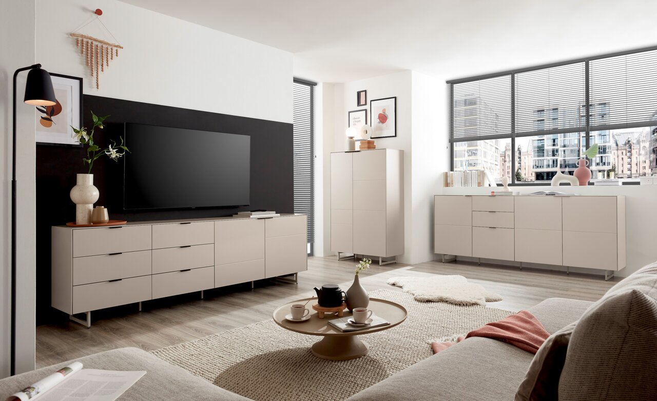 Z2 Dressoir QUODA Moderne woonkamer met lichtbeige dressoirs en een hoog dressoir op metalen poten, een grote tv tegen een donkere accentmuur en een ronde salontafel op een tapijt, gefotografeerd vanuit een iets hoger perspectief vanaf de bank