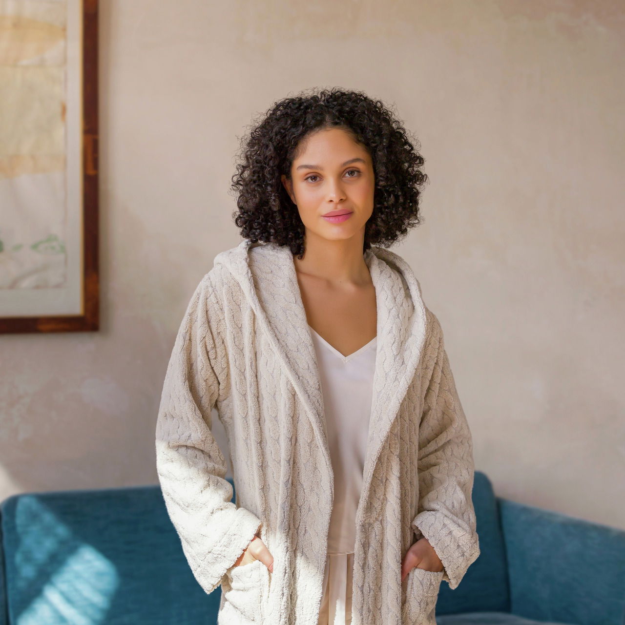 Frau in einem cremefarbenen Kapuzenmantel aus Strick, stehend in einem gemütlichen Raum, von vorne betrachtet.