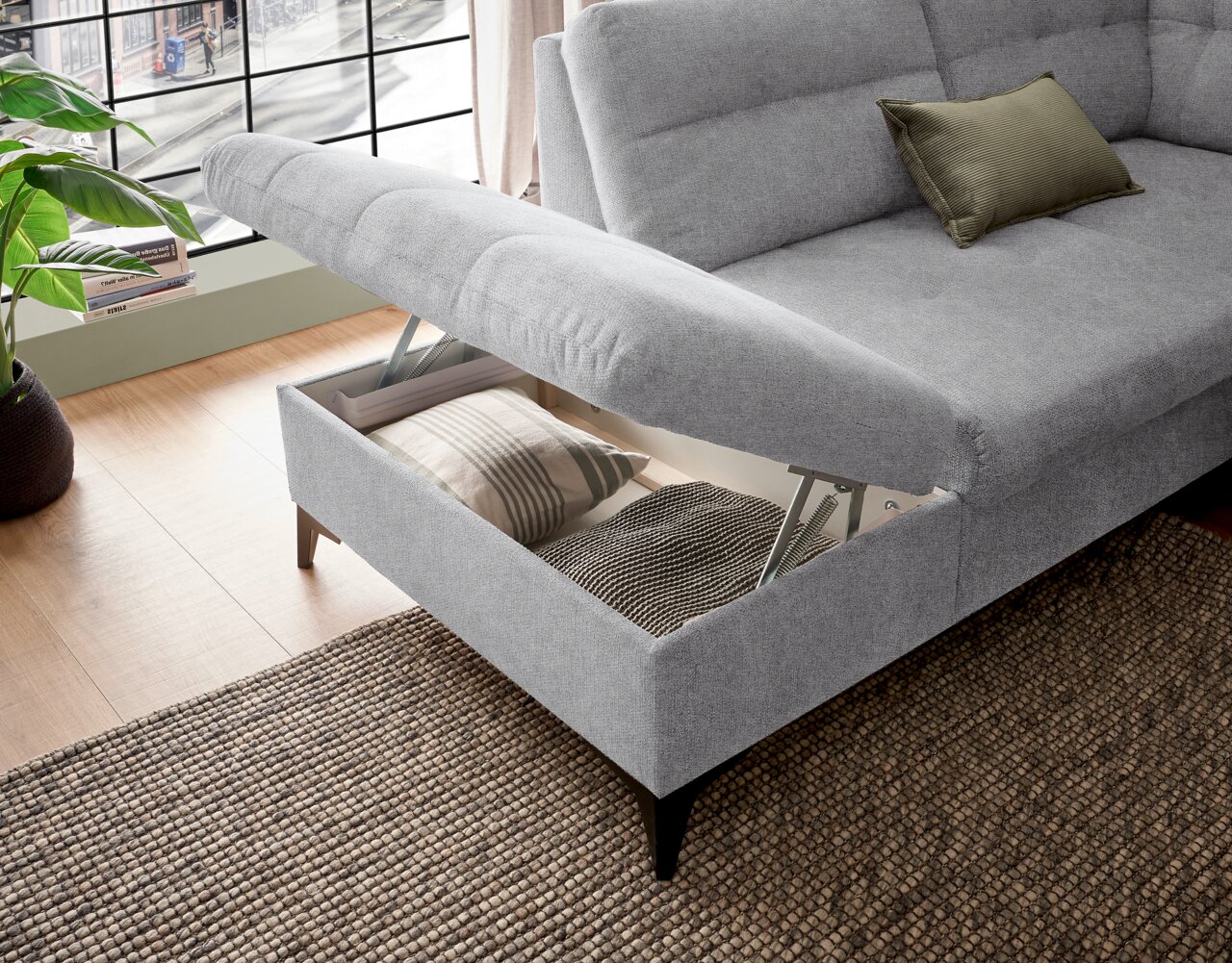 Graues Ecksofa mit Stauraum, geöffnet, von schräg oben fotografiert. Kissen im Stauraum sichtbar.