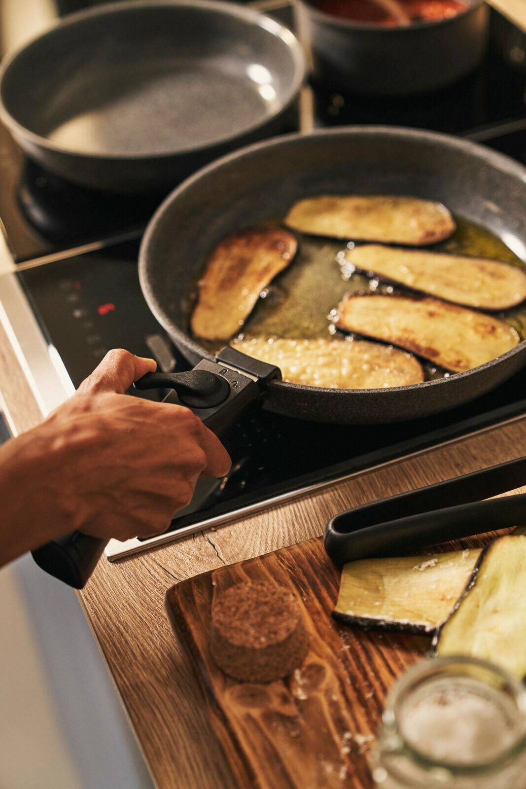 BALLARINI Pfannenset 3-tlg. Nahaufnahme einer Pfanne auf einem Herd, in der Auberginenscheiben gebraten werden. Eine Hand hält den Griff der Pfanne. Im Hintergrund ist eine weitere Pfanne zu sehen.