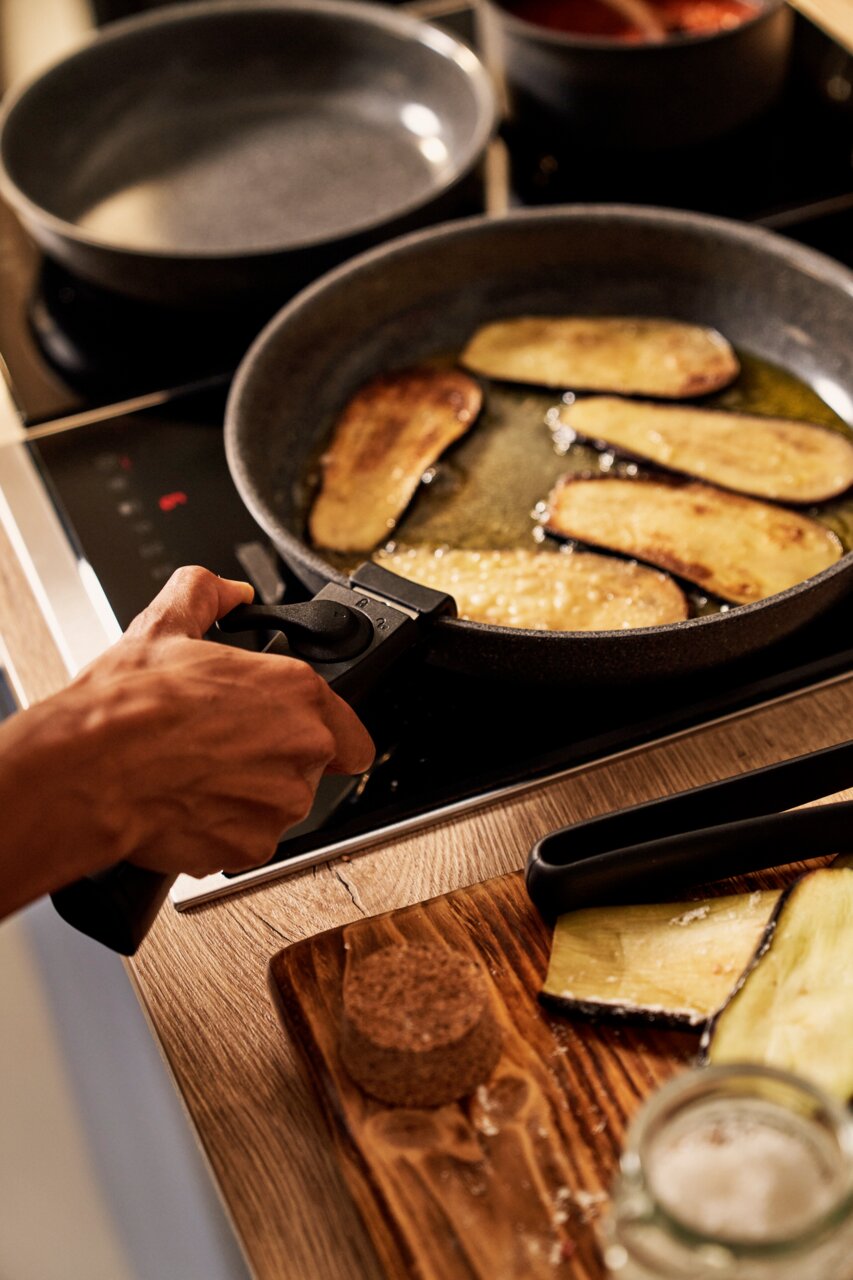 BALLARINI Pfannenset 3-tlg. Nahaufnahme einer Pfanne auf einem Herd, in der Auberginenscheiben gebraten werden. Eine Hand hält den Griff der Pfanne. Im Hintergrund ist eine weitere Pfanne zu sehen.