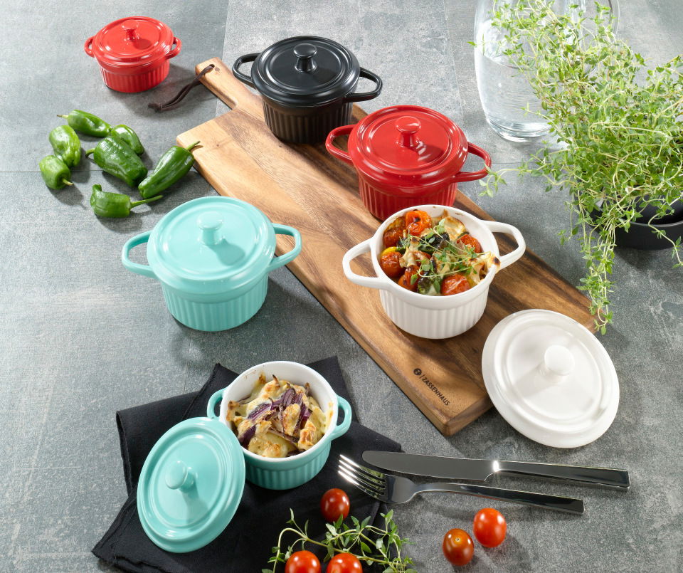 Verschiedene Cocottes in verschiedenen Farben, darunter eine schwarze Cocotte, auf einem Tisch mit Gemüse und Besteck, aus der Vogelperspektive.