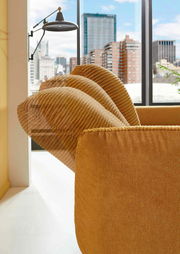 Nahaufnahme einer senfgelben Rundecke mit gerippten Kissen vor einem großen Fenster mit Stadtblick.