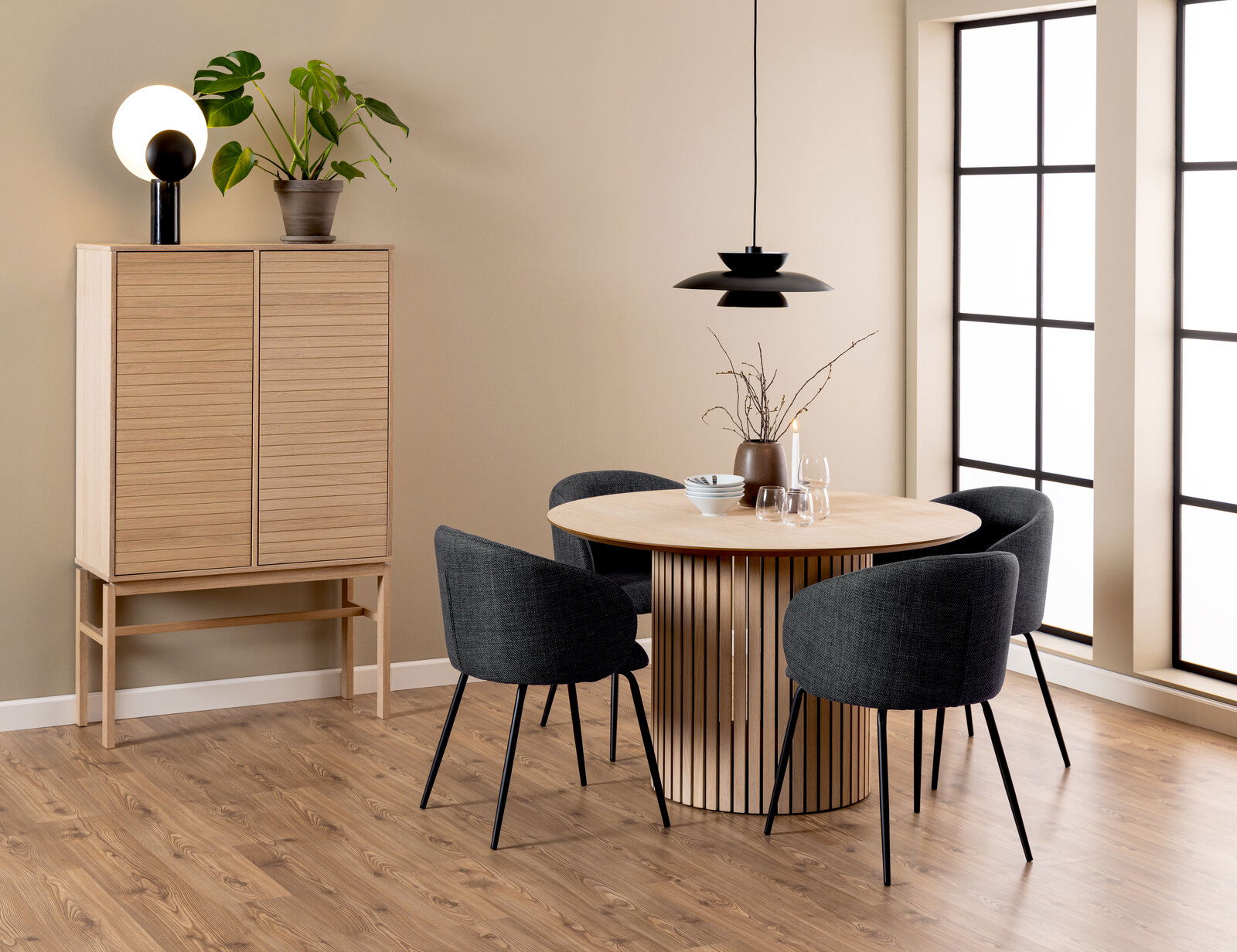 Z2 Esstisch rund LINLEY Moderner Esstisch aus Holz mit runder Tischplatte und vertikalen Streben, umgeben von vier dunkelgrauen Stühlen, in einem stilvoll eingerichteten Raum mit Holzschrank und Pflanzen, seitliche Perspektive.
