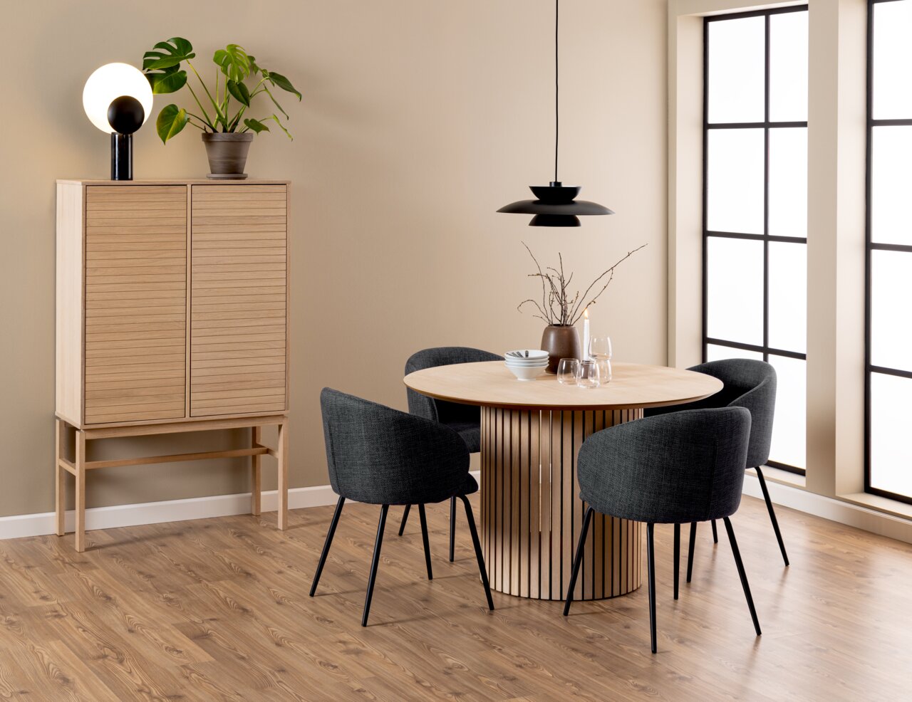 Moderner Esstisch aus Holz mit runder Tischplatte und vertikalen Streben, umgeben von vier dunkelgrauen Stühlen, in einem stilvoll eingerichteten Raum mit Holzschrank und Pflanzen, seitliche Perspektive.