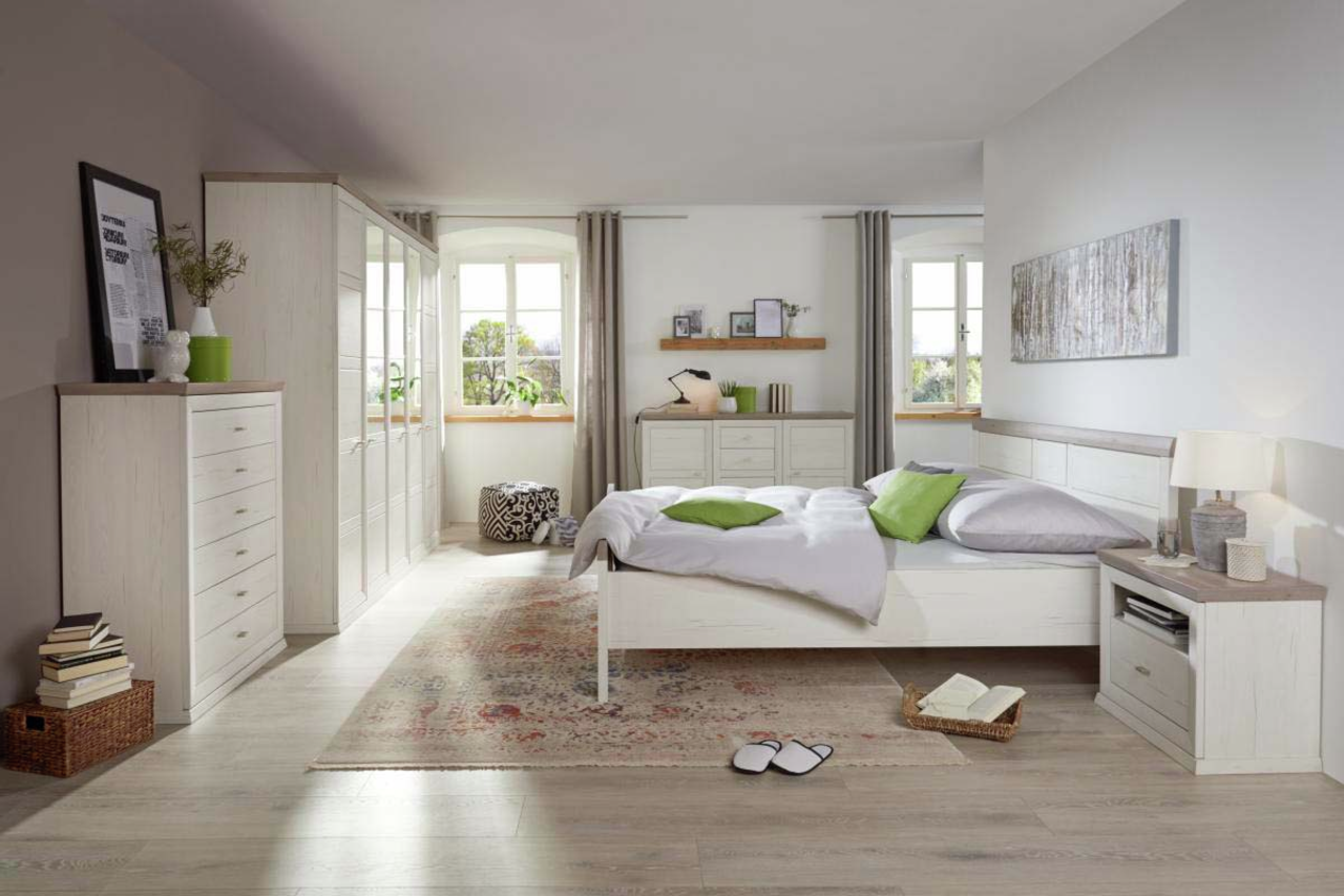 Helles Schlafzimmer mit einem Bett 140x200 in der Mitte, umgeben von weißen Möbeln und dekorativen Elementen, aus der Perspektive von der Zimmertür aus gesehen.