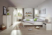 Helles Schlafzimmer mit einem Bett 140x200 in der Mitte, umgeben von weißen Möbeln und dekorativen Elementen, aus der Perspektive von der Zimmertür aus gesehen.