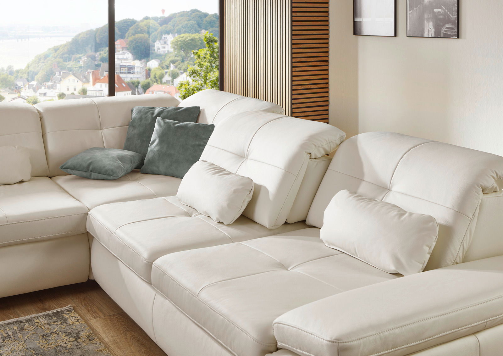Eckgarnitur in cremefarbenem Leder mit mehreren Kissen, fotografiert aus einer seitlichen Perspektive mit Blick auf ein großes Fenster und eine malerische Aussicht.