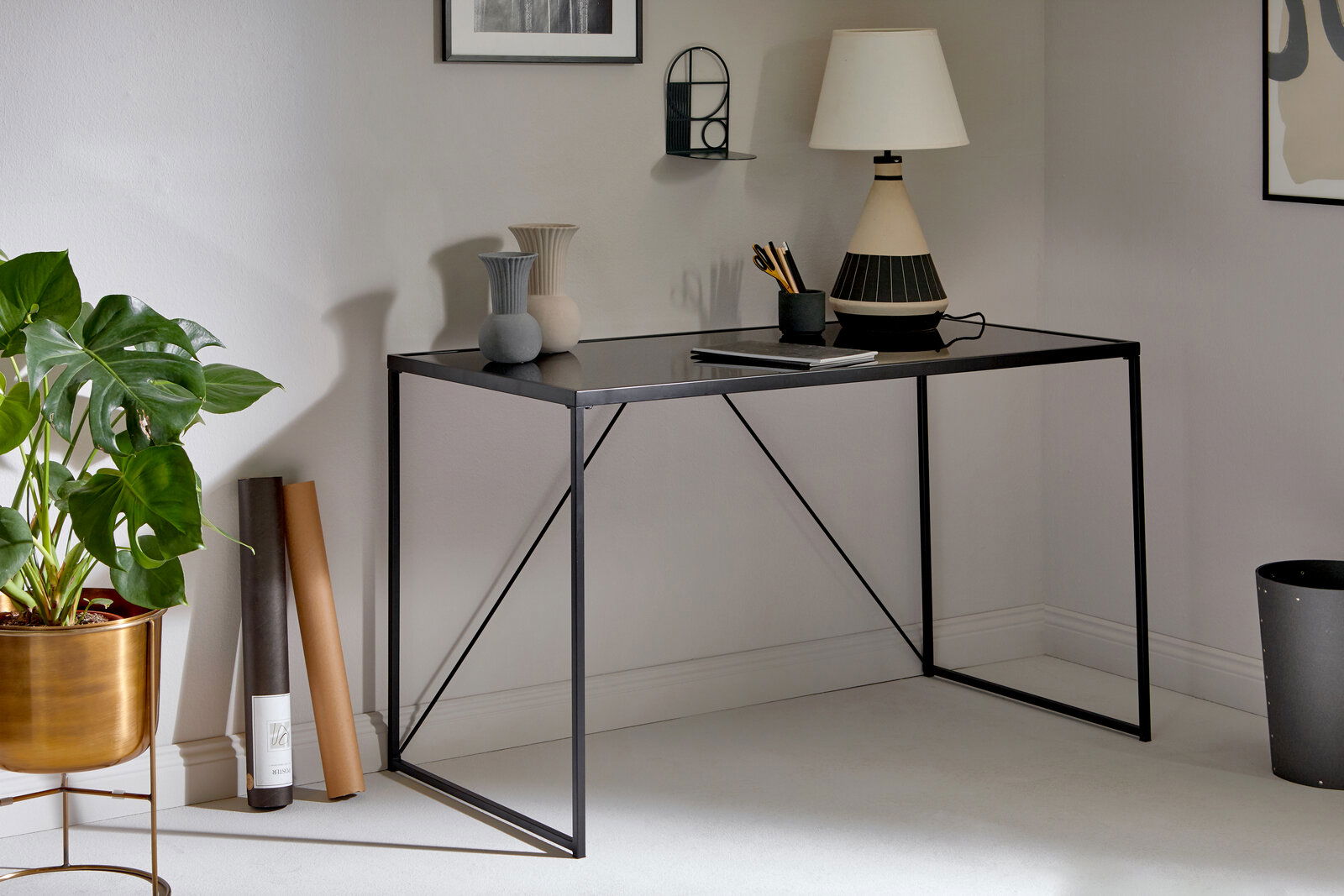 Moderner Schreibtisch mit schwarzer Glasplatte und schlankem Metallgestell, seitliche Perspektive, dekoriert mit Vasen, Lampe und Pflanzen.