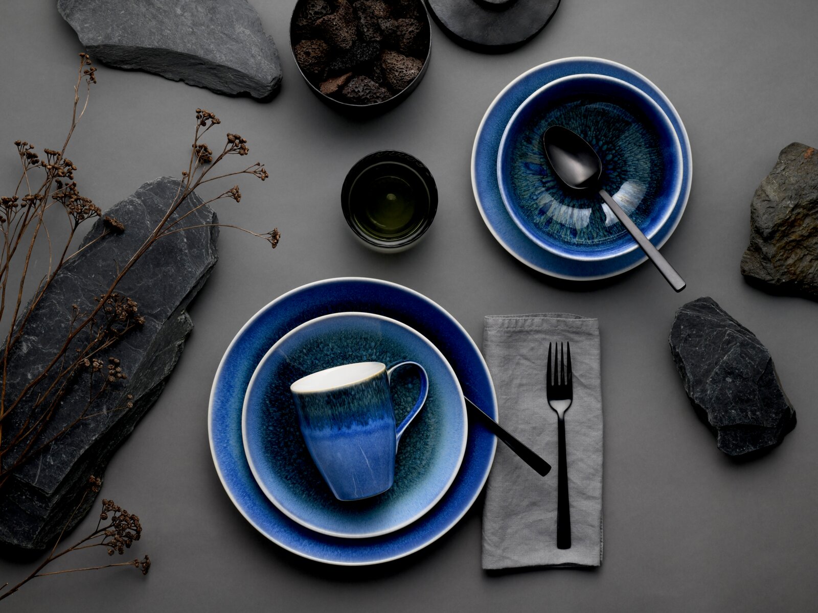Dunkelblau glasiertes Geschirrset mit Tasse auf gestapelten Tellern sowie Schale mit Löffel, grauer Serviette und schwarzem Besteck auf grauem Hintergrund, Draufsicht (Flatlay)