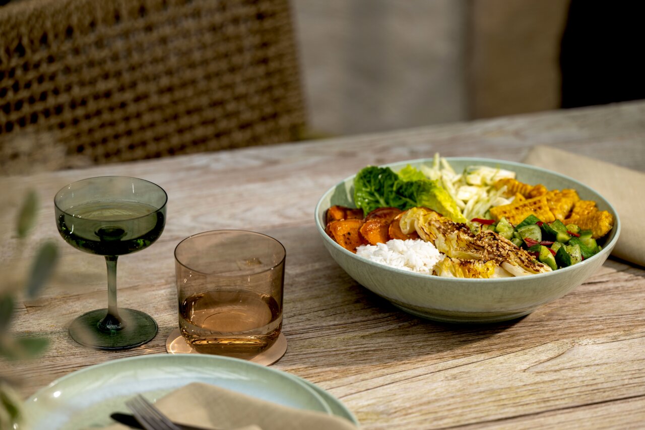 Eine grüne Schale mit einer bunten Auswahl an frischen Zutaten, darunter Reis, Salat, gegrilltes Gemüse und Tempeh, auf einem Holztisch. Die Schale wird aus einer seitlichen Perspektive gezeigt.