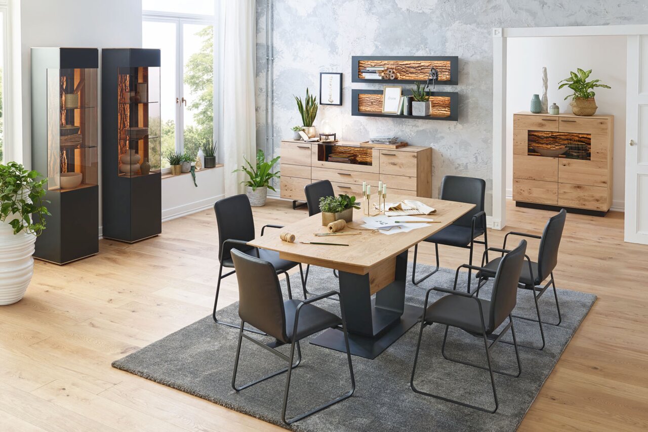Moderner Essbereich mit Esstisch aus Holz und Metall, umgeben von schwarzen Stühlen. Im Hintergrund stehen zwei Vitrinen und ein Sideboard aus Holz. Die Perspektive zeigt den Raum von der Seite mit Blick auf den Tisch und die Möbel im Hintergrund.
