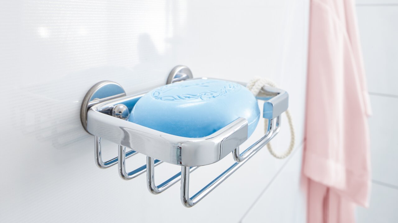 Tesa Seifenkorb ALUXX CHROME Nahaufnahme eines verchromten Seifenkorbs von tesa Aluxx mit blauer Seife, montiert an einer weißen Badezimmerwand.