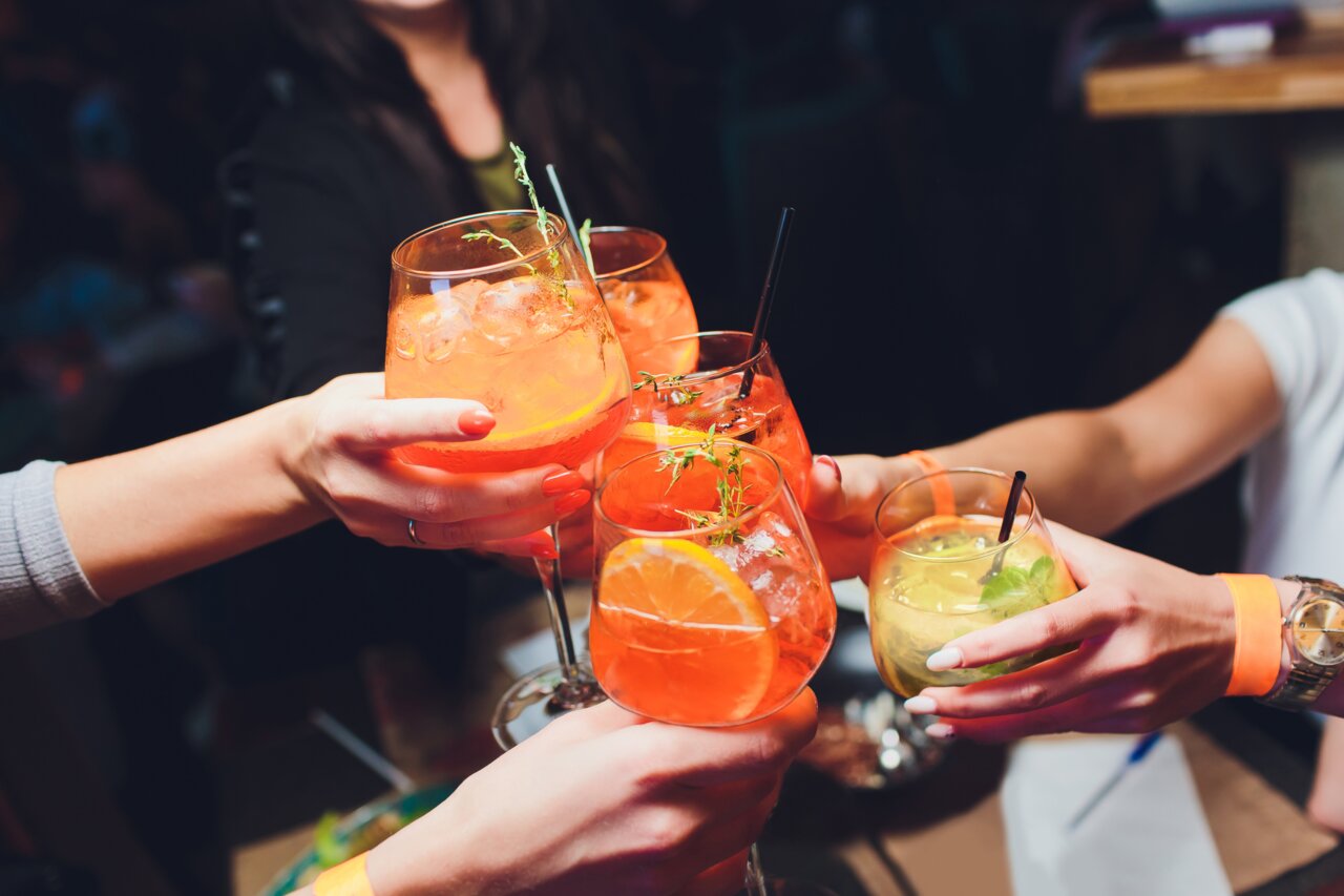 Meerdere mensen proosten met kleurrijke cocktails in ronde glazen, gezien van bovenaf.